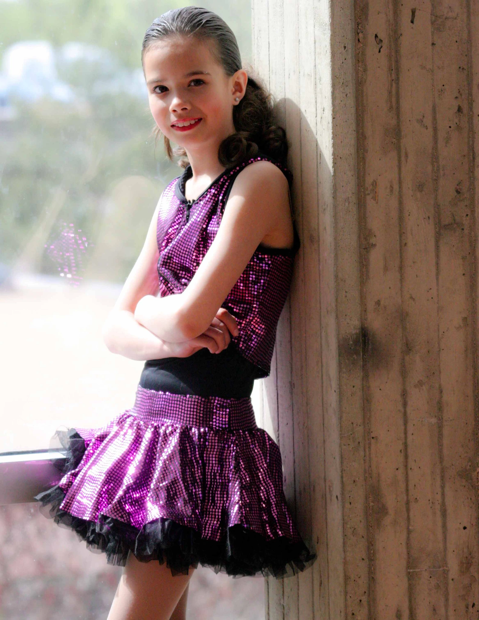 A young dancer in a sequinned costume leaning against a timber post, smiling at the camera
