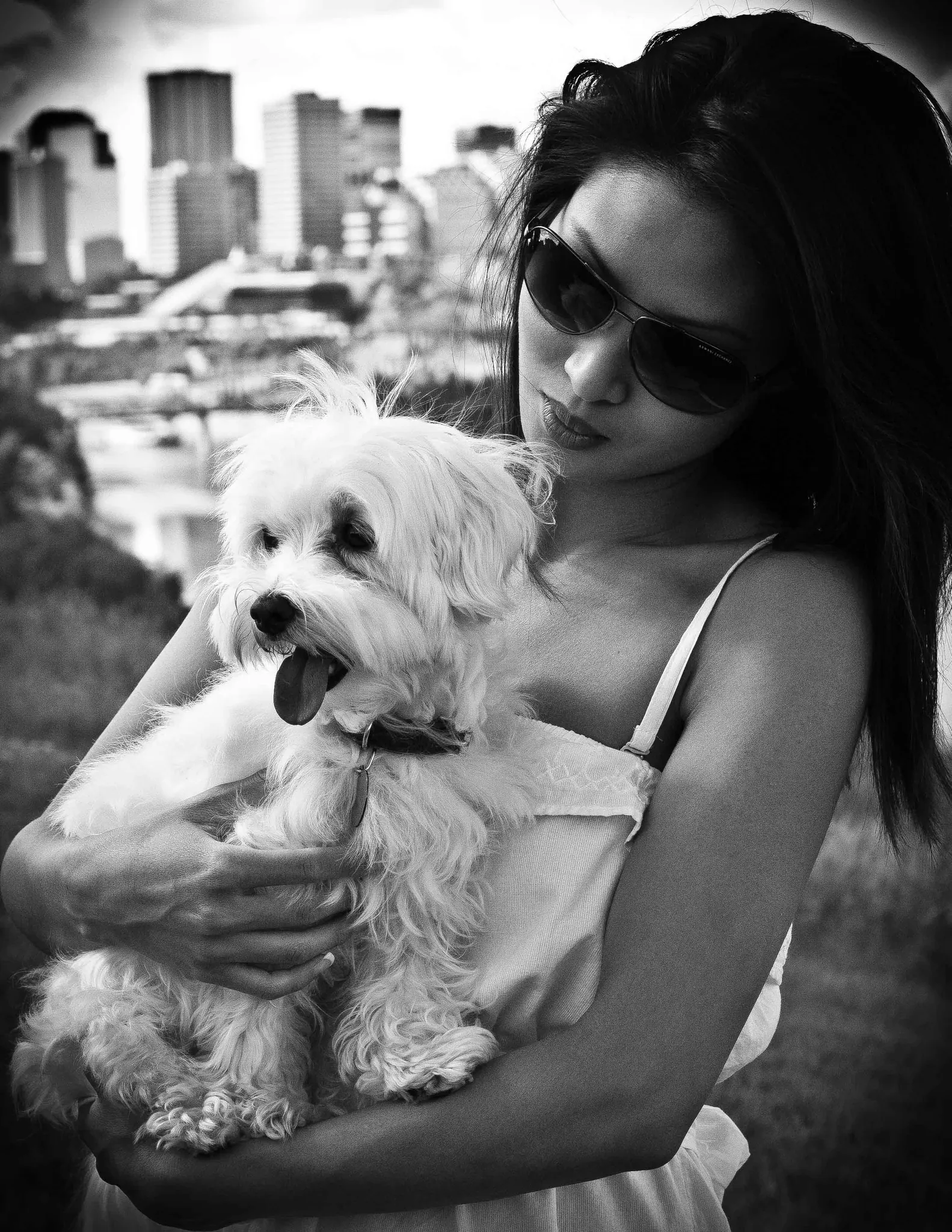 A woman in sunglasses holding a small white dog close, downtown Edmonton behind her, in black and white