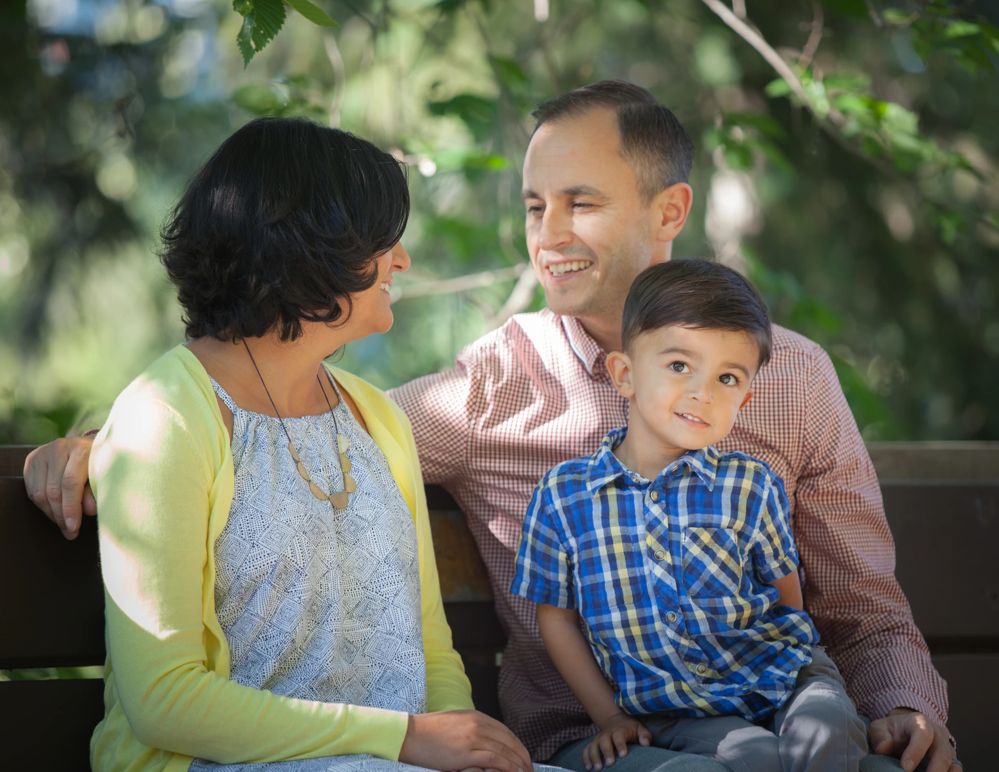 A family of three sitting close together outdoors, the parents laughing with their young son